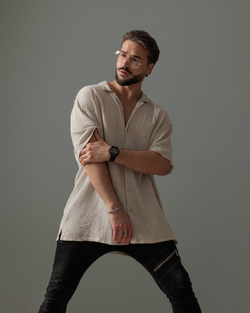 cool fashion man with glasses looking away and holding arms in fashion pose in front of grey backgroundの写真素材