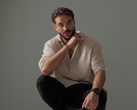 attractive man with glasses crouching with elbows on knees and looking forward in front of grey backgroundの写真素材