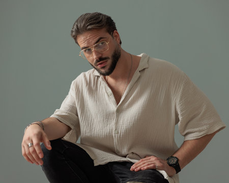 handsome man with glasses looking to side and posing with elbow on knee in front of grey backgroundの写真素材