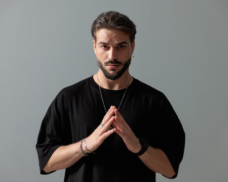 confident unshaved man in black t-shirt touching fingers and posing in front of dark grey backgroundの写真素材