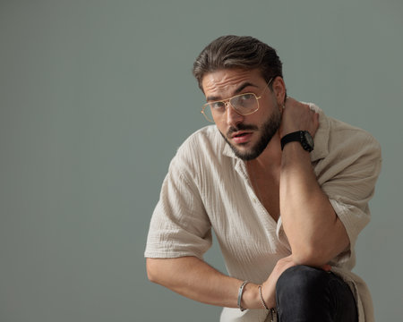 confident young man with glasses holding hand behind neck and posing with elbow on knee in front of grey backgroundの写真素材