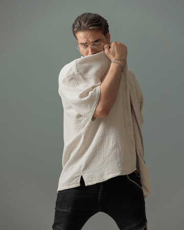 cool young man with glasses covering face with shirt and posing in front of grey backgroundの写真素材