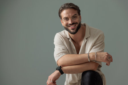 happy young man with glasses holding knee up and folding arms while smiling and looking forward in front of grey backgroundの写真素材