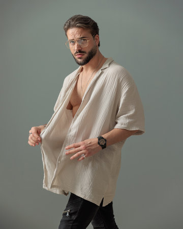 side view of sexy bearded man with glasses arranging and closing shirt while walking in front of grey backgroundの写真素材