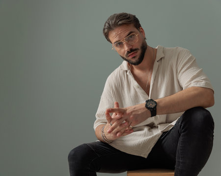 attractive young man with glasses sitting and posing with elbows on knees in front of grey backgroundの写真素材