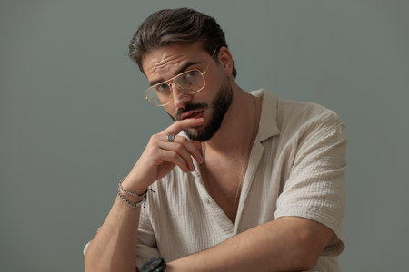 seductive young man with glasses touching lips and looking forward in front of grey backgroundの写真素材