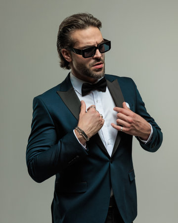 confident elegant businessman with sunglasses looking away and adjusting tuxedo in front of grey backgroundの写真素材