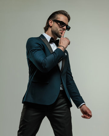 handsome unshaved groom with sunglasses looking to side and making silence gesture while posing in front of grey backgroundの写真素材