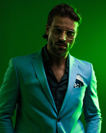 elegant young man with glasses holding hands in pockets and posing in front of green background with green lightsの写真素材