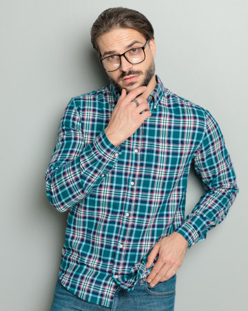 thoughtful casual man in shirt with glasses touching chin and thinking while standing and posing in front of grey backgroundの写真素材