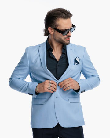 portrait of sexy unshaved guy with sunglasses looking to side while fixing suit in front of white background in studioの写真素材