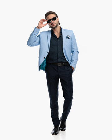 unshaved elegant man in suit with hand in pocket adjusting sunglasses and moving in front of white backgroundの写真素材