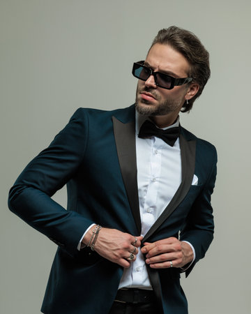 portrait of bearded young man with sunglasses looking to side and adjusting tuxedo while confidently posing on grey backgroundの写真素材