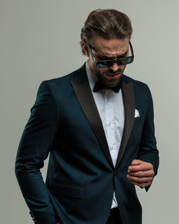 elegant young groom with sunglasses looking down and holding hands in pockets in front of grey backgroundの写真素材