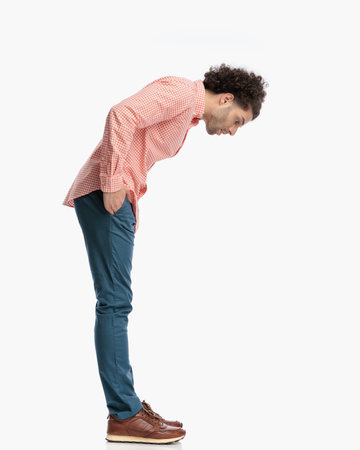 curly hair man with hands in pockets leaning forward and looking down trying to see something small on the floor in front of white backgroundの写真素材