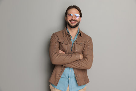happy young man with glasses crossing arms and laughing in front of grey background in studioの写真素材