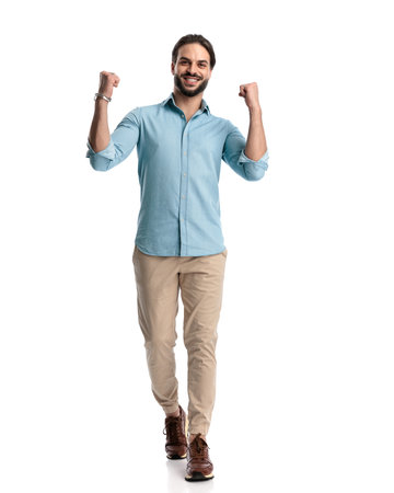 excited casual man with beard holding fists up and celebrating victory, walking in front of white background in studioの写真素材