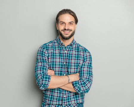 portrait of proud casual man in plaid shirt crossing arms and smiling in front of grey backgroundの写真素材