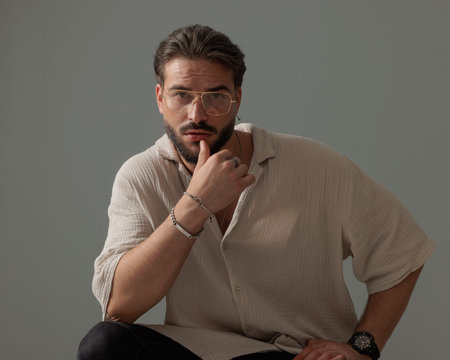 bearded young man with glasses sensually touching lips and crouching with elbow on knee in front of grey backgroundの写真素材