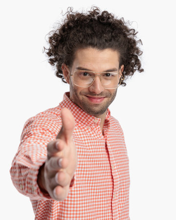 happy young man with curly hair wearing glasses and smiling while shaking hand in front of white background in studioの写真素材