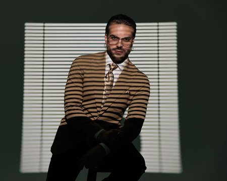 sexy elegant businessman with glasses holding elbows on knees, looking forward and sitting in front of window shadow on grey backgroundの写真素材