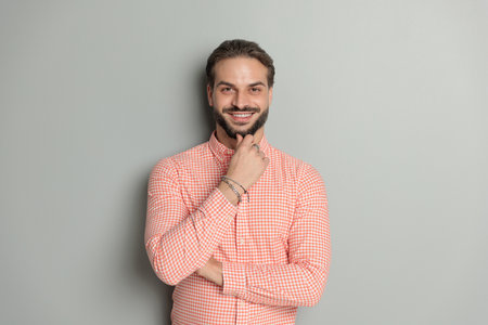 thoughtful happy young man folding arms, touching beard and thinking while smiling in front of grey backgroundの写真素材