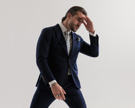 thoughtful young man holding hand to forehead and looking away while wearing elegant navy blue suit with tie and handkerhchief on grey backgroundの写真素材