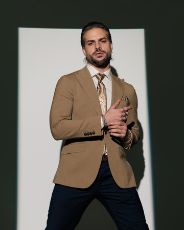 dramatic cool man in luxury brown suit holding and touching hands in a fashion pose while standing in front of window shadow on grey backgroundの写真素材