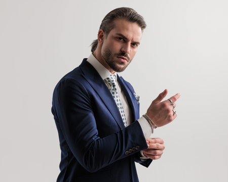 side view of elegant high class businessman in navy blue suit with tie and handkerchief adjusting sleeves and looking forward on grey backgroundの写真素材