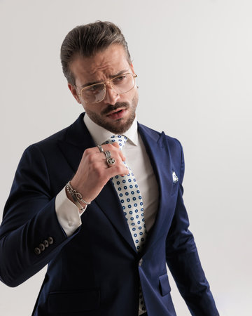 sexy high class businessman in navy blue suit looking away while adjusting tie in front of grey backgroundの写真素材