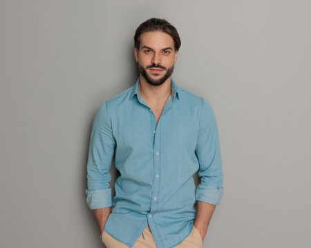young gasual guy with beard holding hands in pockets and looking forward in front of grey backgroundの写真素材