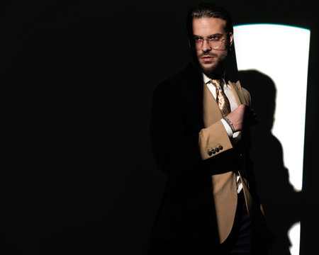 high class elegant man with glasses looking away and holding arms in fashion pose and posing in front of window shadow on grey backgroundの写真素材