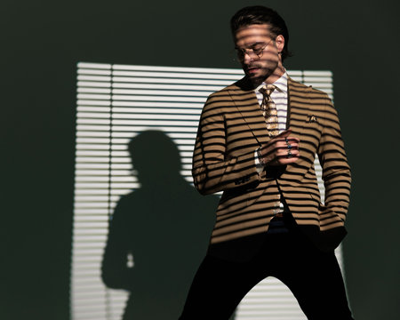 elegant young businessman with glasses looking down and posing with one hand in pocket while holding arm in a cool way in front of window shadow on grey backgroundの写真素材