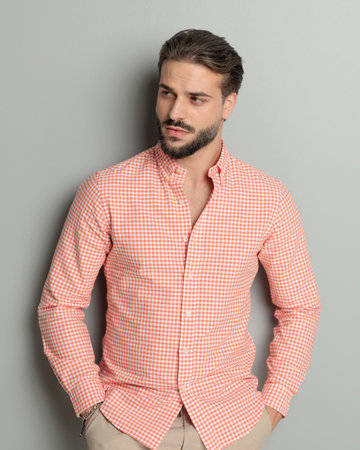 sexy young man with beard holding hands in pockets and looking to side while confidently posing on grey backgroundの写真素材