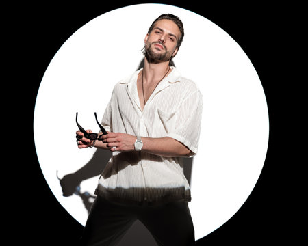 sexy cool man holding sunglasses, feeling confident and posing in front of round spotlight on white backgroundの写真素材