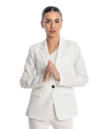 serious young woman in white suit holding and touching palms while looking forward in front of white backgroundの写真素材
