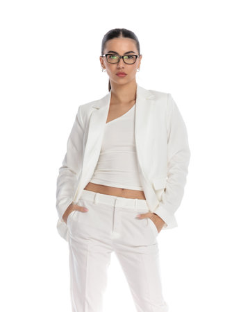 sexy young girl student with glasses wearing white suit, holding hands in pockets and looking forward in front of white backgroundの写真素材
