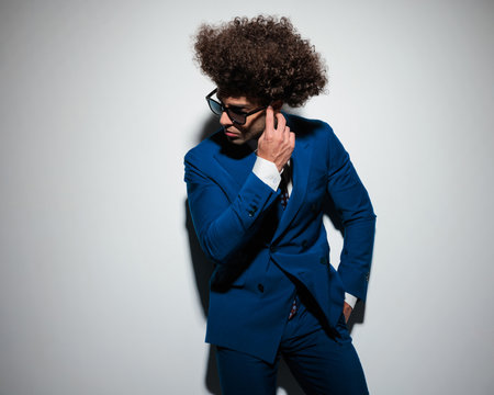 sexy high class businessman with long curly hair looking to side and adjusting sunglasses, holding hand in pocket and posing on grey backgroundの写真素材