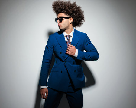 cool fashion man with sunglasses looking to side and adjusting blue suit in front of grey backgroundの写真素材