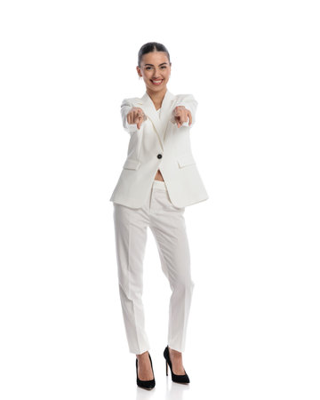 excited elegant young woman in white suit smiling while pointing fingers forward in front of white background in studioの写真素材