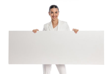 happy beautiful woman in white suit showing empty board and smiling in front of white backgroundの写真素材
