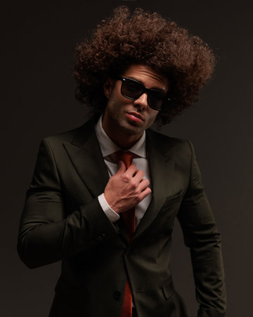 sexy young man with long curly hair and sunglasses adjusting tie and posing in front of grey backgroundの写真素材
