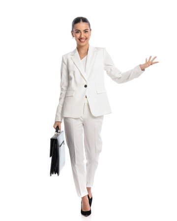 confident young lady in white suit with bag showing and inviting to side while walking in front of white backgroundの写真素材