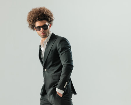 portrait of elegant young man with glasses wearing open collar shirt with black tuxedo and holding hands in pockets on grey backgroundの写真素材