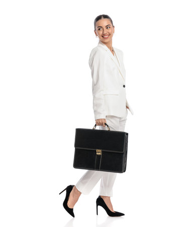 side view of happy elegant woman with bag looking behind and smiling while walking in front of white backgroundの写真素材