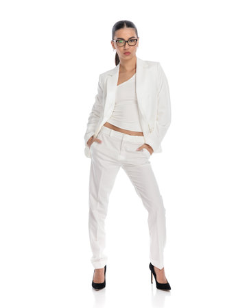 elegant young businessman in white suit with glasses holding hands in pockets and posing in a confident way on white backgroundの写真素材