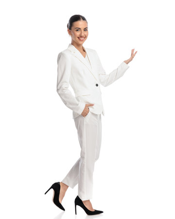 proud young woman in white suit showing to side with one hand while holding the other in pocket and stepping on white backgroundの写真素材