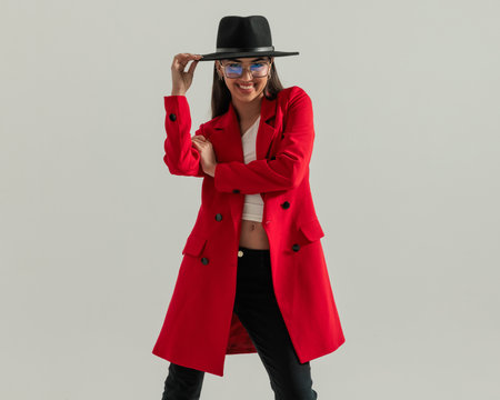 happy casual woman with glasses adjusting hat and smiling while wearing red coat in front of grey backgroundの写真素材