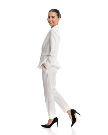 side view of elegant young businesswoman in white suit being happy, smiling and walking with hands in pockets in front of white backgroundの写真素材