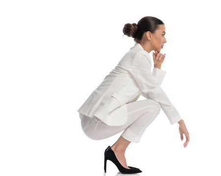 full body picture of elegant young woman in white suit touching chin and thinking while holding elbows on knees and crouching on white backgroundの写真素材